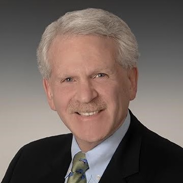 Mark Mandell wearing a dark suit and tie in front of a grey background.