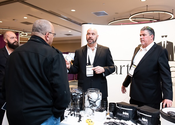 Three men in dark suits talking at an exhibit booth.